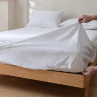 A person’s hands are shown fitting a white sheet onto a mattress on a wooden bed frame, with white pillows and another sheet already on the bed.