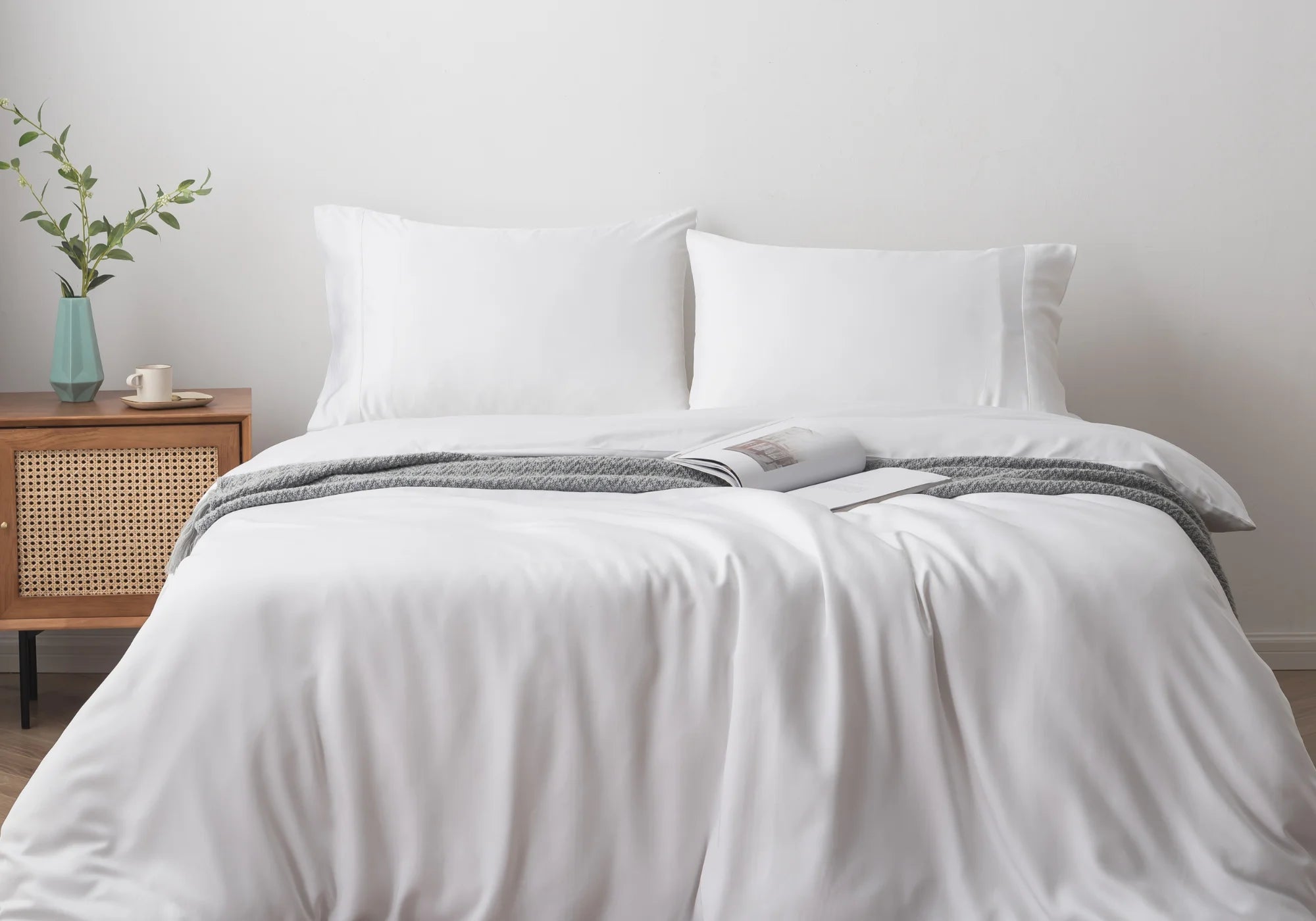 A neatly made bed with white bedding, two pillows, and a gray throw blanket sits against a white wall. An open magazine lies on the bed. A wooden nightstand with a plant and vase is on the left.