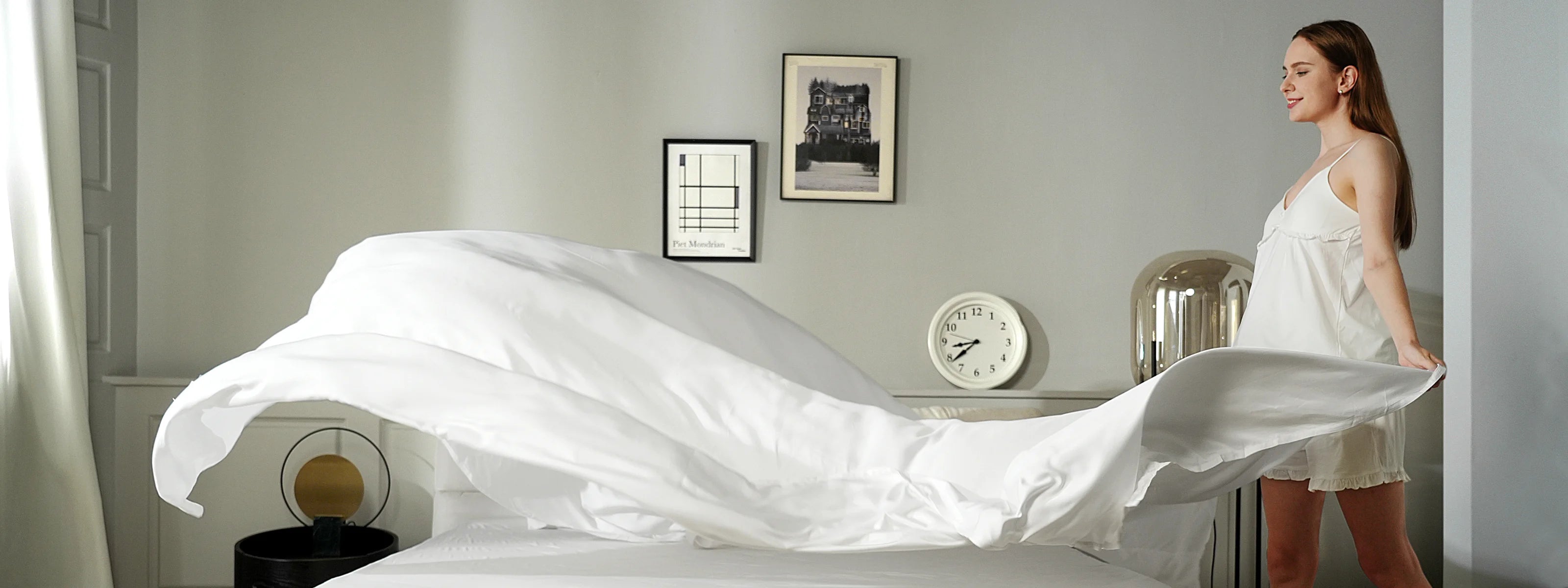 A woman in a white nightgown is making a bed, spreading a white sheet over it in a bright, modern bedroom with framed pictures and a clock on the wall.