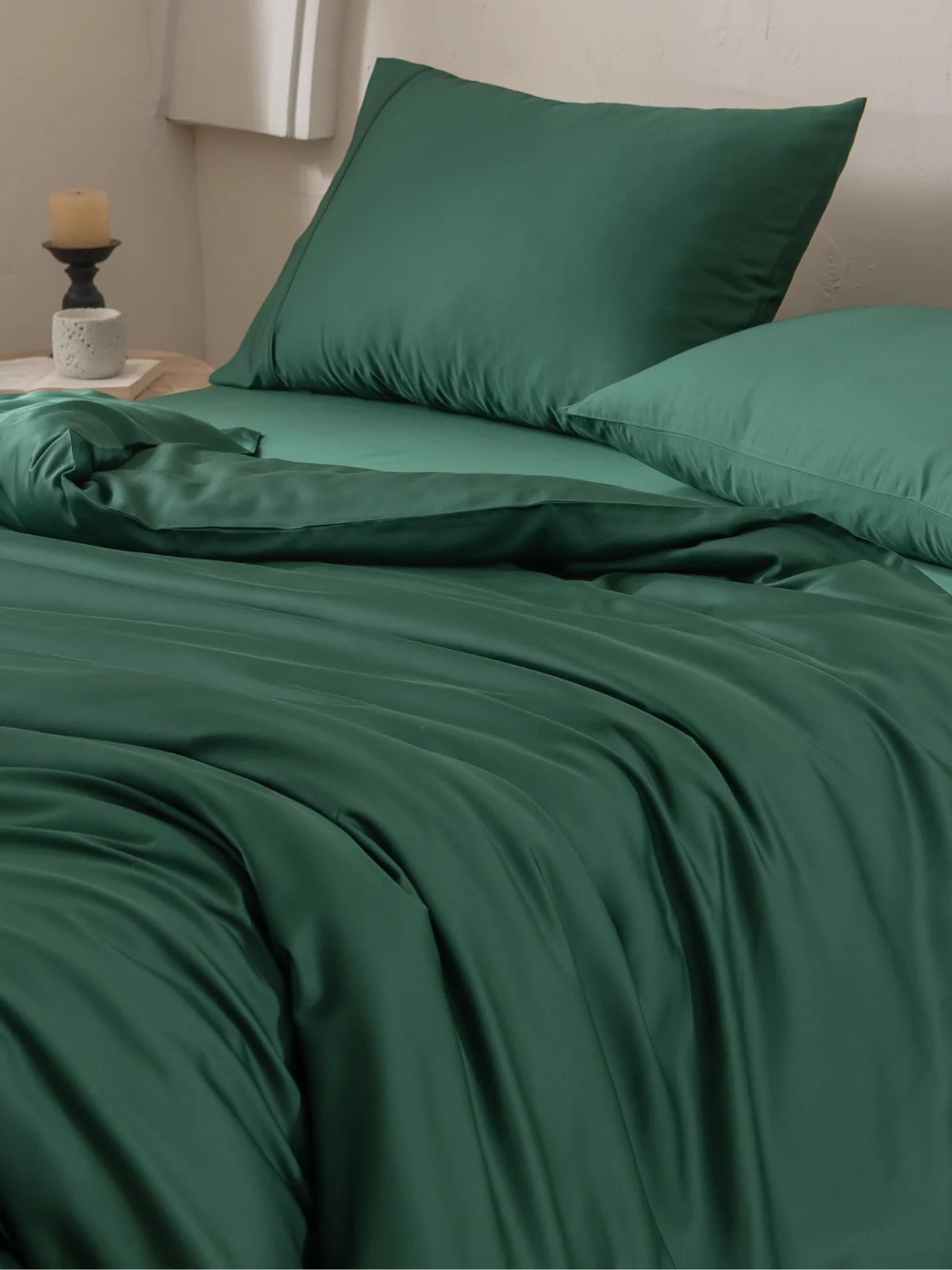 A neatly made bed with dark green sheets and pillowcases, next to a wooden nightstand holding a lit candle and a small white vase, set against a light-colored wall.