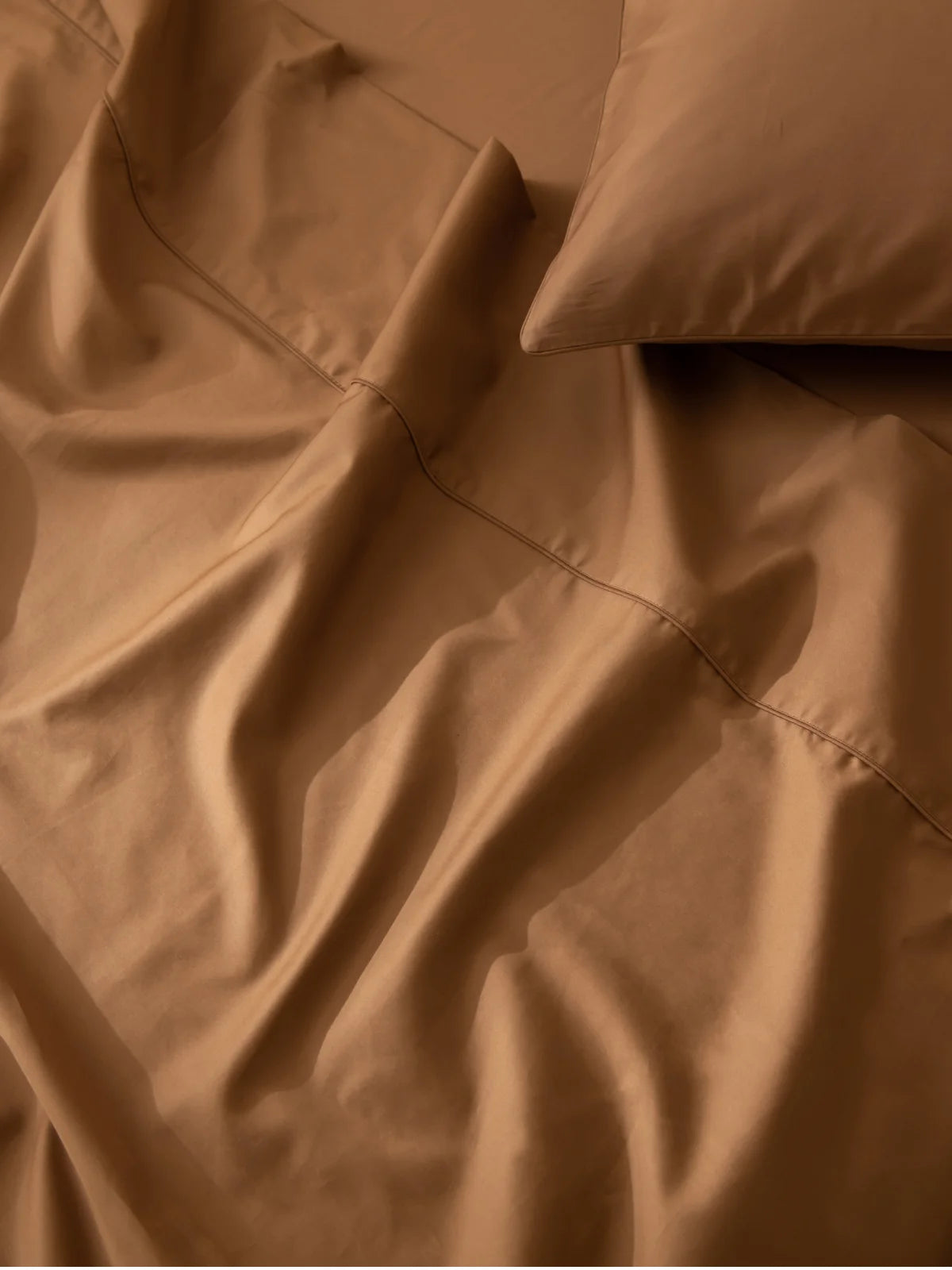 A close-up view of a neatly made bed with smooth, brown sheets and a matching pillowcase, showing some gentle folds and soft lighting.