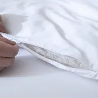 A hand holds open the zipper of a white pillowcase, revealing the pillow insert inside. The background is also white, creating a clean and minimal look.