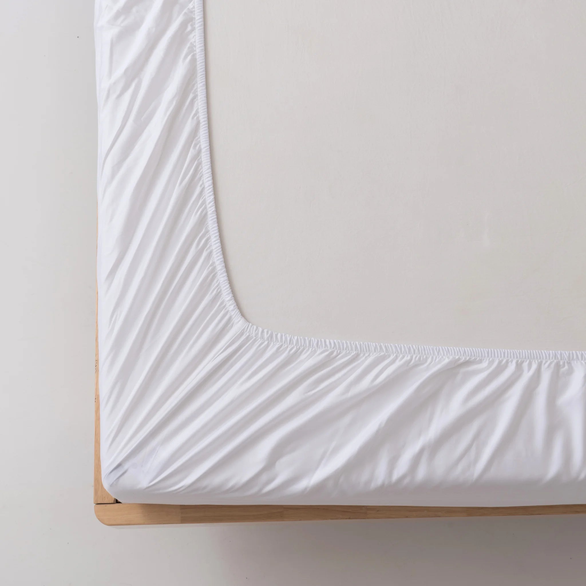 A close-up of a neatly made bed corner with the Linenly Bamboo Fitted Sheet - White, featuring deep sides snugly wrapped around a mattress on a light wooden bed frame.