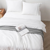 A neatly made bed features the Linenly Bamboo Quilt Cover Set - White in a soft sateen weave, with white sheets and pillows, a gray patterned throw, and an open book on top. A wooden nightstand sits beside the bed on a light-colored floor.
