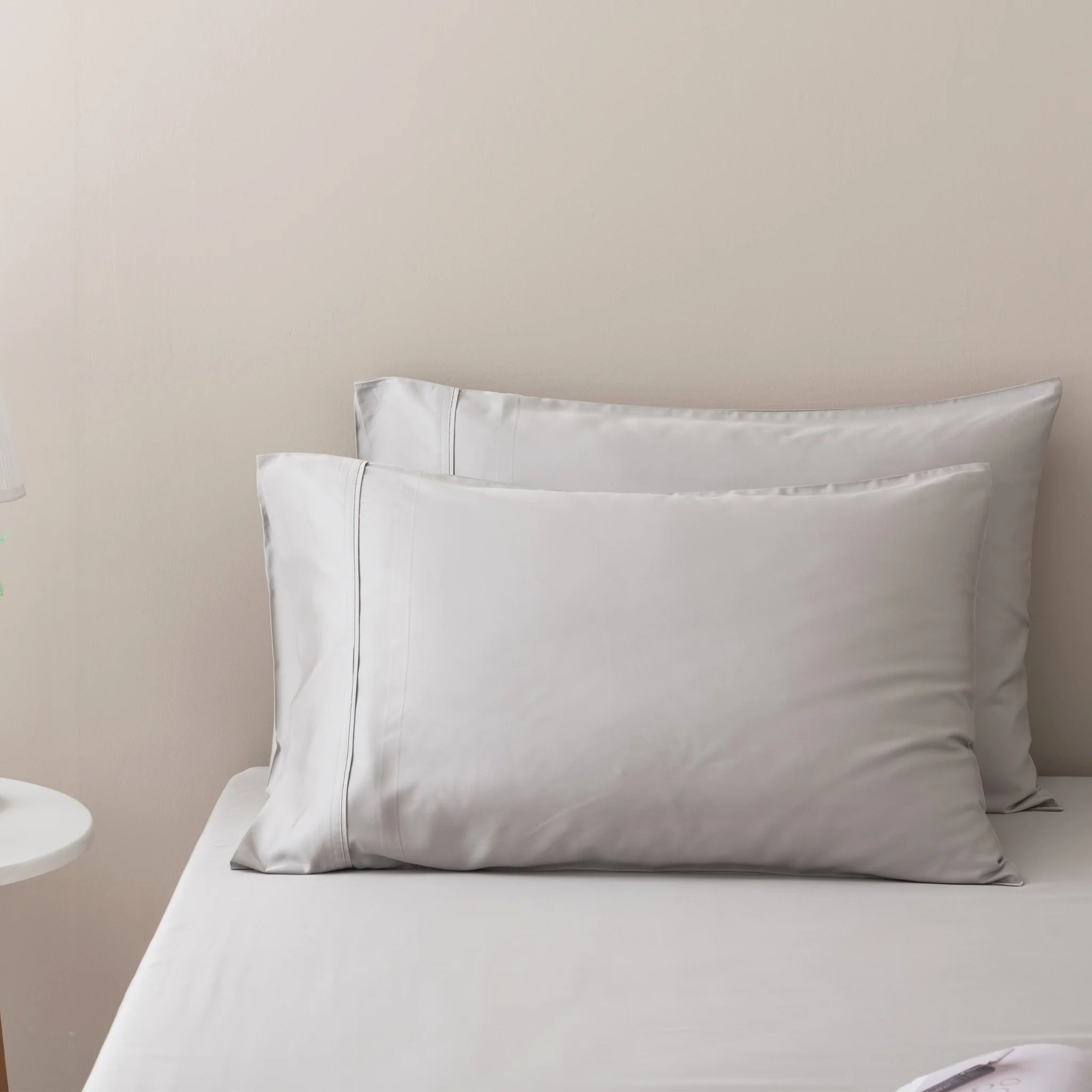 Two neatly arranged white pillows rest against a light wall on a made bed dressed in Linenly’s Bamboo Sheet Set - Silver; a small round bedside table is partially visible on the left.