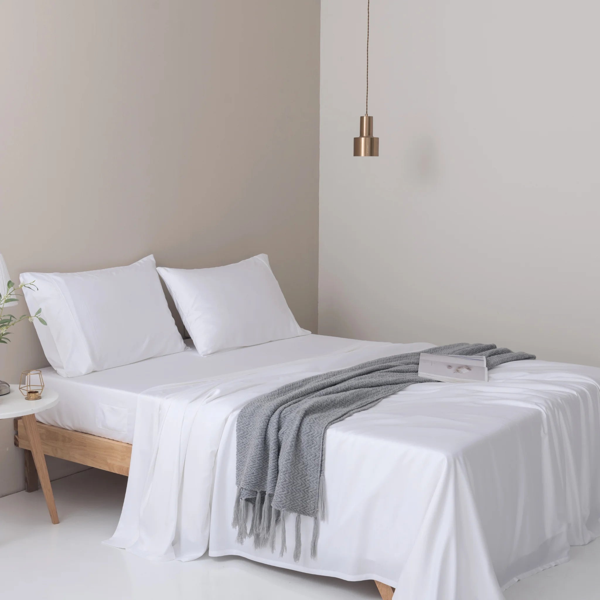 A neatly made bed is dressed in ultra-soft, breathable Linenly Bamboo Sheet Set - White, with two pillows and a gray throw. An open book rests on the bed, while a small side table holds a plant and candle beneath a modern pendant light.