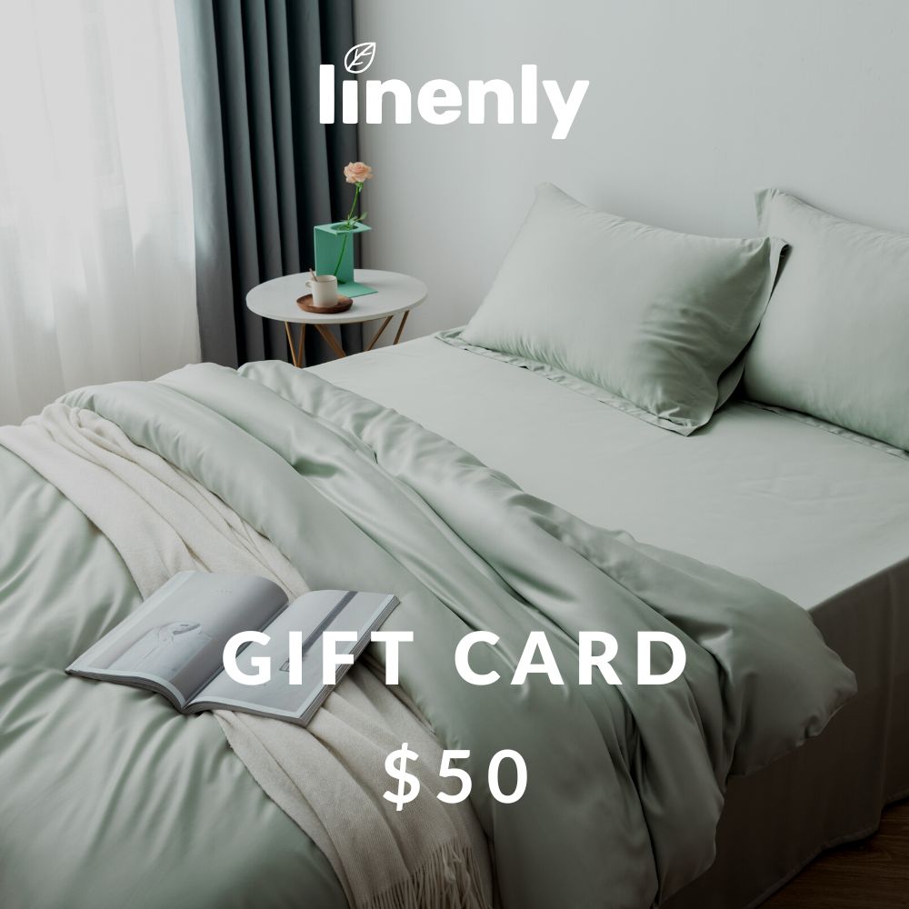 A neatly made bed with light green sheets and pillows is shown, accompanied by a cozy white blanket. On the bed, there is an open book. A small round side table holds a cactus plant. The image features text: "Linenly Gift Cards," perfect for promoting a good night's sleep or digital gift cards.